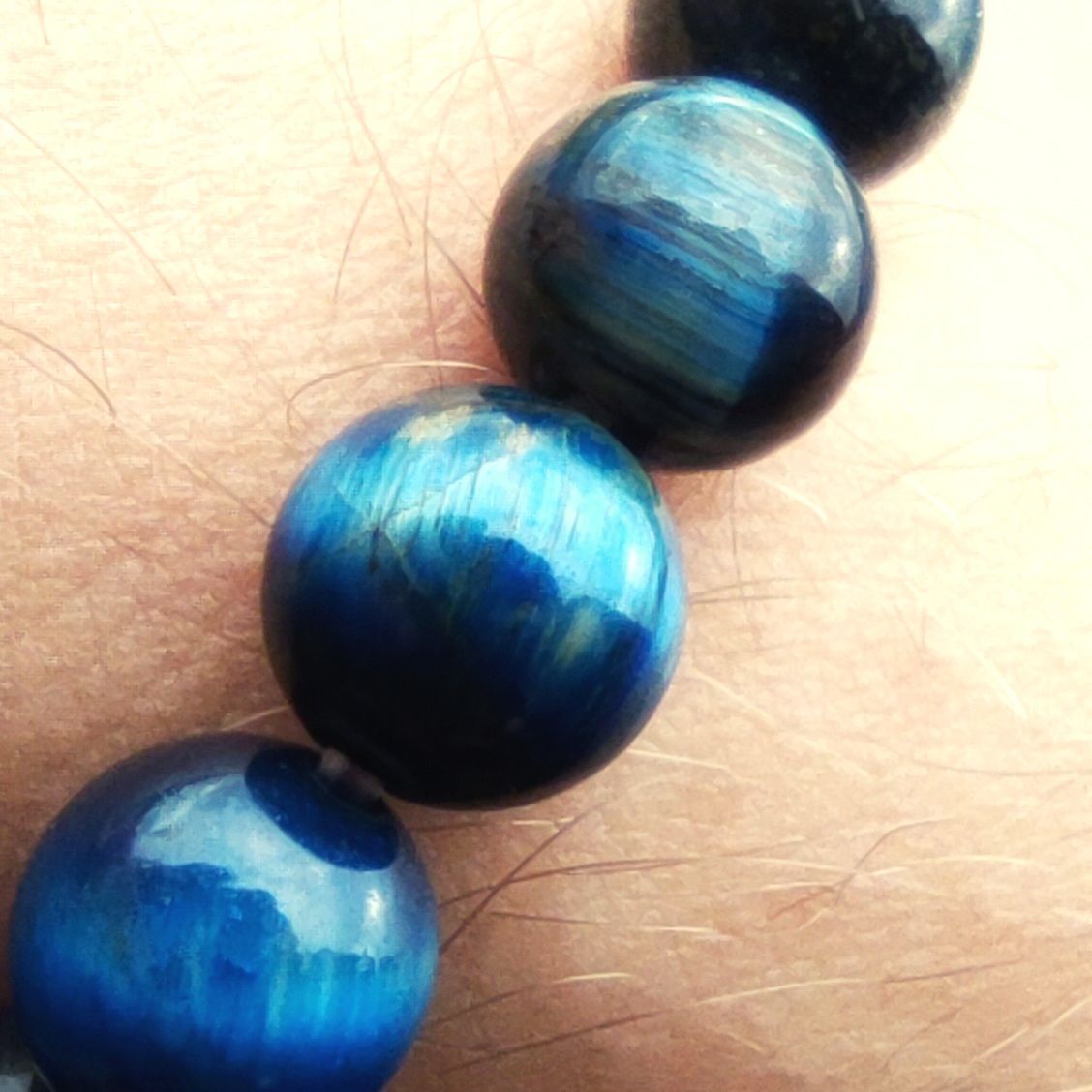 Macro close-up detail of natural blue Tiger Eye beads on skin, showing the stone's silky chatoyant texture and deep blue hues.
